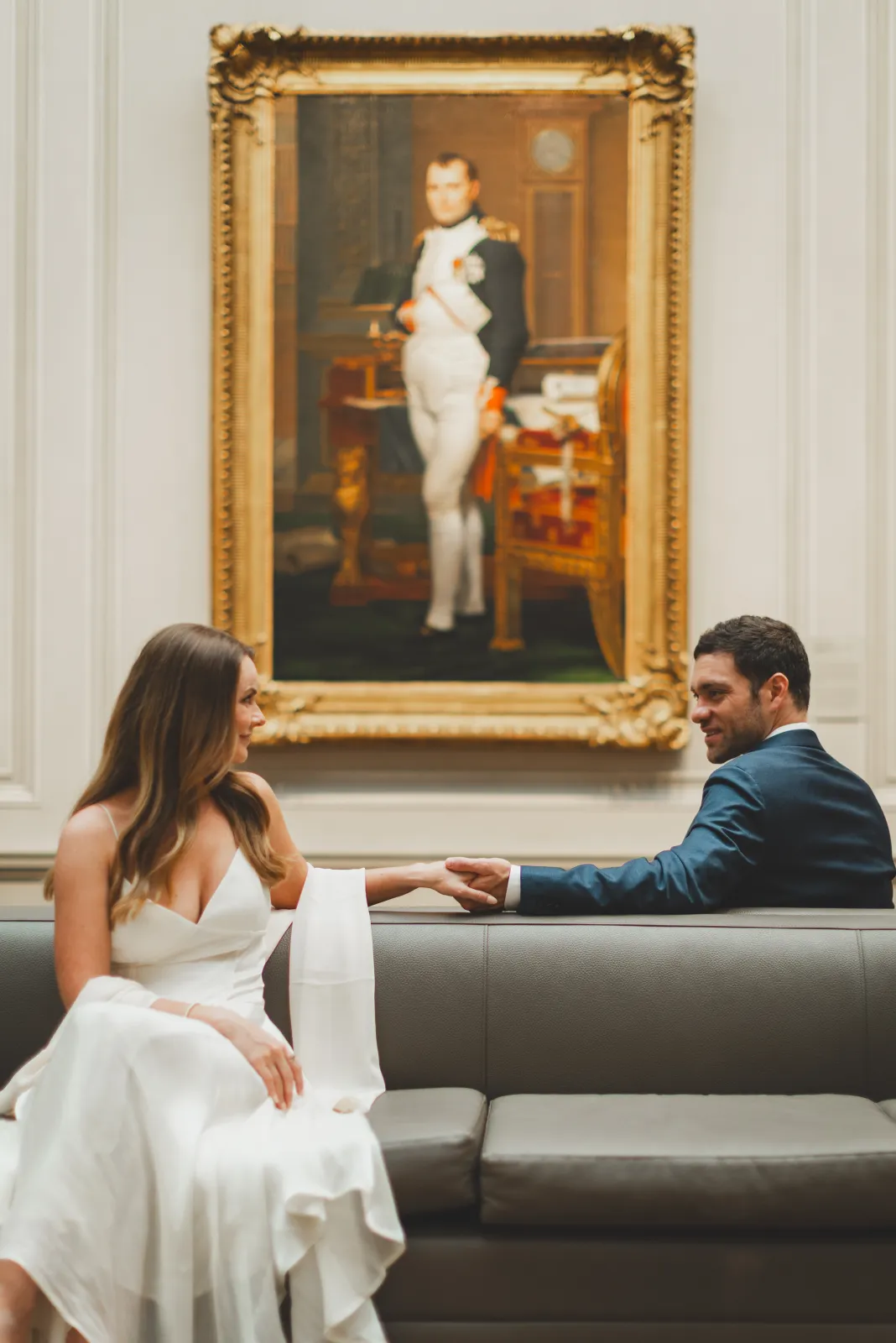 Holding hands on a gallery bench beneath a portrait of Napoleon